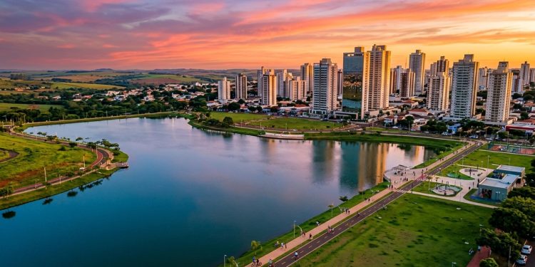 Vista panorâmica de cidade brasileira ao pôr do sol, com lago urbano, prédios, áreas verdes e ciclovia ao redor.