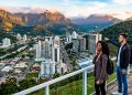 Casal observa cidade cercada por montanhas em mirante panorâmico, com prédios, rio, áreas verdes e paisagem urbana ao entardecer.