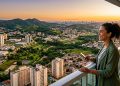 Mulher observa cidade arborizada do alto de uma varanda, com prédios, bairros residenciais, áreas verdes e pôr do sol ao fundo.
