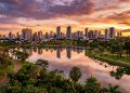 Vista panorâmica de cidade com prédios refletidos em lago, parque arborizado, pistas de caminhada e céu colorido ao pôr do sol.