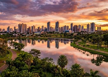 Vista panorâmica de cidade com prédios refletidos em lago, parque arborizado, pistas de caminhada e céu colorido ao pôr do sol.