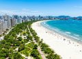 Vista aérea da orla de Santos, com faixa de areia extensa, mar azul, prédios altos e jardins à beira-mar.