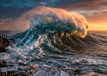 Onda gigante quebra próxima a rochas costeiras ao pôr do sol, com espuma intensa, mar agitado e pessoas observando ao longe.