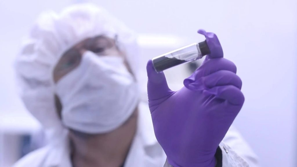 Pesquisadora em laboratório usa máscara, touca e luvas roxas enquanto segura um pequeno tubo com amostra escura.