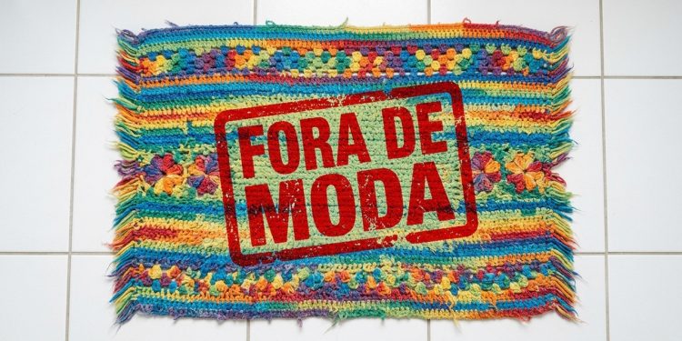 Tapete de crochê colorido sobre piso branco aparece com carimbo vermelho com a frase “Fora de moda”.