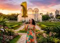 Mulher segura balão dourado em formato de número 1 em jardim florido, com estufa de vidro e paisagem urbana ao fundo.