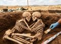 Dois esqueletos humanos aparecem lado a lado em uma escavação arqueológica, posicionados frente a frente em uma sepultura antiga.