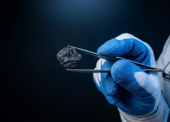 Pessoa com luvas azuis segura um pequeno fragmento escuro de rocha espacial com pinças, em contexto de pesquisa da NASA.
