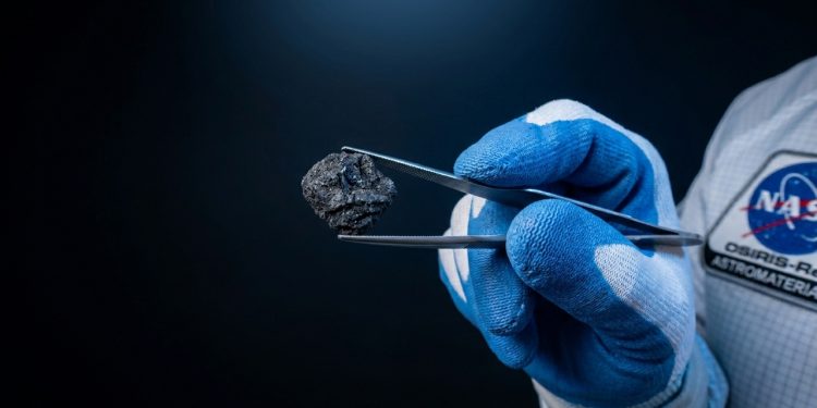 Pessoa com luvas azuis segura um pequeno fragmento escuro de rocha espacial com pinças, em contexto de pesquisa da NASA.