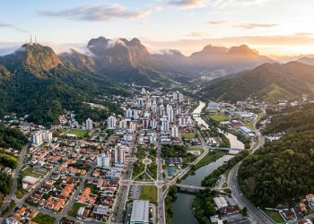 Vista aérea de Jaraguá do Sul, em Santa Catarina, com área urbana cercada por montanhas, mata verde e rio ao entardecer.
