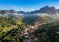 Vista aérea de uma pequena cidade em vale verde, cercada por montanhas, mata preservada, névoa baixa e uma igreja no centro do povoado.