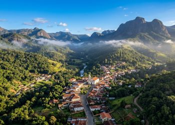 Vista aérea de uma pequena cidade em vale verde, cercada por montanhas, mata preservada, névoa baixa e uma igreja no centro do povoado.