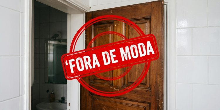 Porta de madeira antiga em banheiro com azulejos brancos, marcada por selo vermelho com a frase “Fora de moda”.