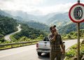 Motorista observa placa de trânsito com símbolo de pneu com correntes em estrada de serra, ao lado de uma caminhonete em paisagem montanhosa.