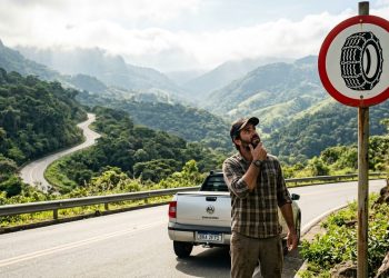Motorista observa placa de trânsito com símbolo de pneu com correntes em estrada de serra, ao lado de uma caminhonete em paisagem montanhosa.