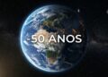 Visão realista do planeta Terra flutuando no espaço sideral, mostrando o continente africano e europeu iluminados pelas luzes das cidades durante a noite. Bem no centro, letras grandes e brancas formam o texto "-50 ANOS".