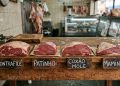 Peças frescas de carne bovina alinhadas sobre uma grossa mesa de madeira rústica em um açougue. À frente de cada peça, uma placa de madeira indica o corte: Contrafilé, Patinho, Coxão Mole e Maminha. Carnes penduradas aparecem desfocadas ao fundo.