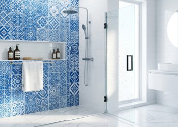 Banheiro moderno com box de vidro, chuveiro, azulejos azuis decorativos e bancada branca com espelho.