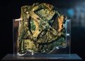 Fragmento do Mecanismo de Anticítera em exposição, com engrenagens antigas de bronze corroído sobre suporte transparente.