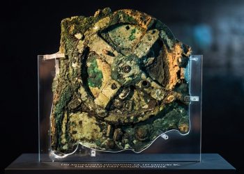 Fragmento do Mecanismo de Anticítera em exposição, com engrenagens antigas de bronze corroído sobre suporte transparente.
