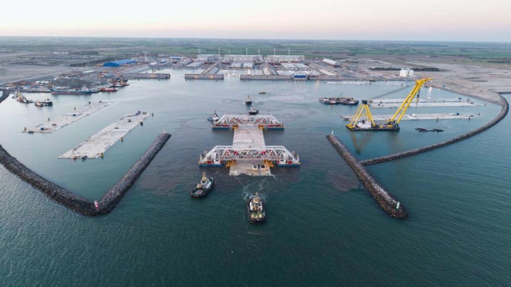 Vista aérea de uma obra marítima com guindaste, rebocadores e estruturas de concreto sendo movimentadas no mar.