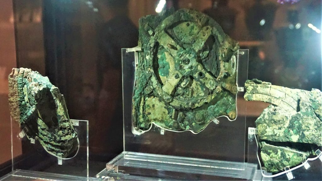 Fragmentos do Mecanismo de Anticítera expostos em museu, com engrenagens antigas preservadas em bronze corroído.