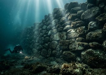 Mergulhador observa uma muralha de pedras submersa, iluminada por feixes de luz dentro do mar.