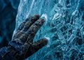 Mão com luva toca uma parede de gelo azul, com cristais e neve acumulada na superfície.