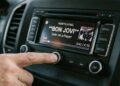 Mão ajustando o botão de volume no painel multimídia de um carro. A tela digital do rádio está acesa e exibe a reprodução da música "Livin' on a Prayer" da banda Bon Jovi.