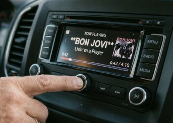 Mão ajustando o botão de volume no painel multimídia de um carro. A tela digital do rádio está acesa e exibe a reprodução da música "Livin' on a Prayer" da banda Bon Jovi.
