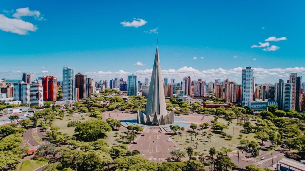 Vista aérea de Maringá com a Catedral Basílica Menor Nossa Senhora da Glória ao centro, cercada por áreas verdes e prédios urbanos.