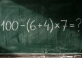 Quadro escolar com a expressão matemática 100 - (6 + 4) x 7 = ? escrita em giz branco.