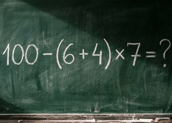 Quadro escolar com a expressão matemática 100 - (6 + 4) x 7 = ? escrita em giz branco.