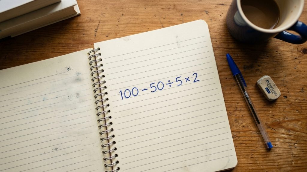 Caderno aberto sobre mesa de madeira com a expressão matemática 100 - 50 ÷ 5 × 2 escrita em caneta azul.