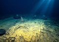 Fundo do oceano em área profunda mostra camadas rochosas fraturadas, sedimentos, escuridão azul e feixes de luz atravessando a água.