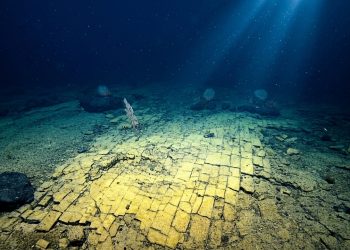 Fundo do oceano em área profunda mostra camadas rochosas fraturadas, sedimentos, escuridão azul e feixes de luz atravessando a água.