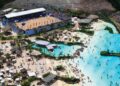 Vista aérea de um grande parque aquático mostrando uma extensa piscina de ondas azuis com dezenas de banhistas, uma praia artificial com espreguiçadeiras e, à esquerda, uma arena esportiva de areia com arquibancadas lotadas.