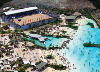 Vista aérea de um grande parque aquático mostrando uma extensa piscina de ondas azuis com dezenas de banhistas, uma praia artificial com espreguiçadeiras e, à esquerda, uma arena esportiva de areia com arquibancadas lotadas.