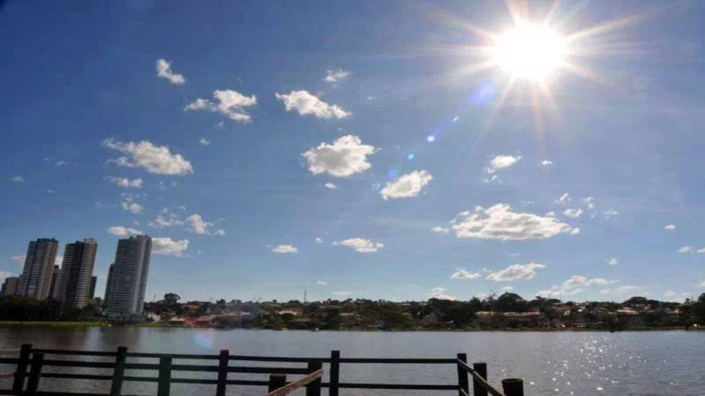 Vista do Parque das Nações Indígenas, com lago, céu ensolarado, prédios ao fundo e área de observação à beira da água.