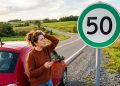 Motorista confusa observa placa de velocidade máxima de 50 km/h ao lado de carro vermelho em estrada rural.
