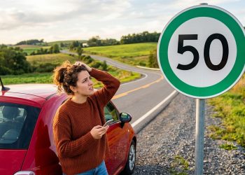 Motorista confusa observa placa de velocidade máxima de 50 km/h ao lado de carro vermelho em estrada rural.