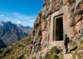 Pessoa observa portal de pedra esculpido em uma parede rochosa nos Andes, com montanhas ao fundo.