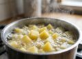 Panela de aço inox sobre o fogão com água em forte ebulição, cozinhando vários cubos amarelos e uniformes de batata, com bastante vapor subindo pela cozinha.