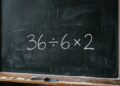 Quadro-negro escolar tradicional com moldura de madeira apresentando a expressão matemática 36 ÷ 6 x 2 escrita centralizada com giz branco. Na base de madeira, encontram-se gizes coloridos e um apagador.