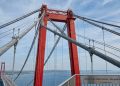 Visão a partir da passarela da Ponte 1915 Çanakkale olhando para cima, destacando a imponente torre de aço vermelho e a complexa rede de grossos cabos de suspensão. No canto inferior direito, um guarda-corpo exibe uma placa com a inscrição: 'Vão central: 2.023 metros - Recorde Mundial'.