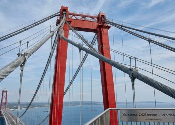 Visão a partir da passarela da Ponte 1915 Çanakkale olhando para cima, destacando a imponente torre de aço vermelho e a complexa rede de grossos cabos de suspensão. No canto inferior direito, um guarda-corpo exibe uma placa com a inscrição: 'Vão central: 2.023 metros - Recorde Mundial'.