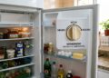 Visão interna de uma geladeira abastecida com diversos potes e alimentos, destacando uma grande ilustração sobreposta na porta mostrando o botão do termostato ajustado na posição "Médio".