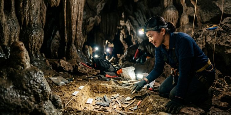 Pesquisadora realiza escavação arqueológica dentro de caverna, com ferramentas, lanternas e vestígios no solo.