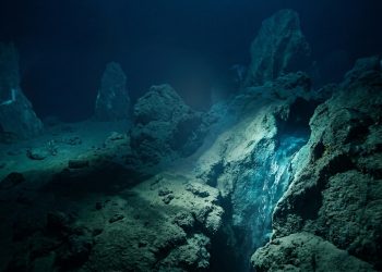 Fenda no fundo do oceano em ambiente escuro, cercada por formações rochosas, sedimentos e luz azulada em águas profundas.