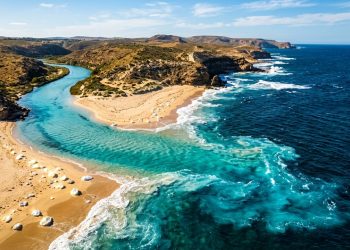 Vista aérea de rio encontrando o oceano em praia de águas azul-turquesa, com falésias, areia clara e paisagem costeira preservada.